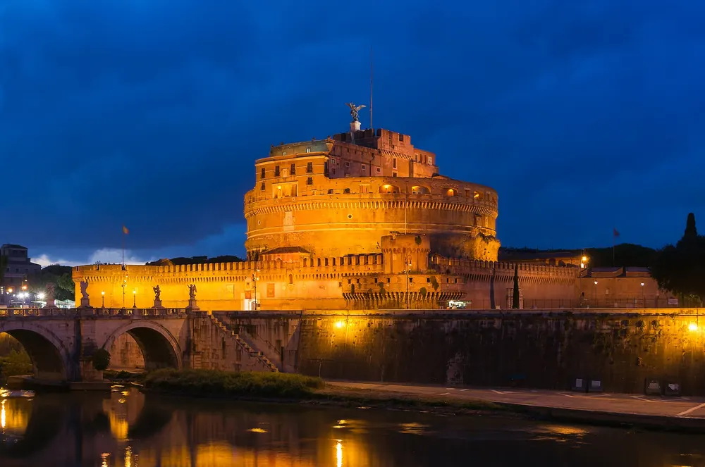 Castel Sant'Angelo and the Ponte Sant'Angelo lit warmly against a deep blue evening sky.