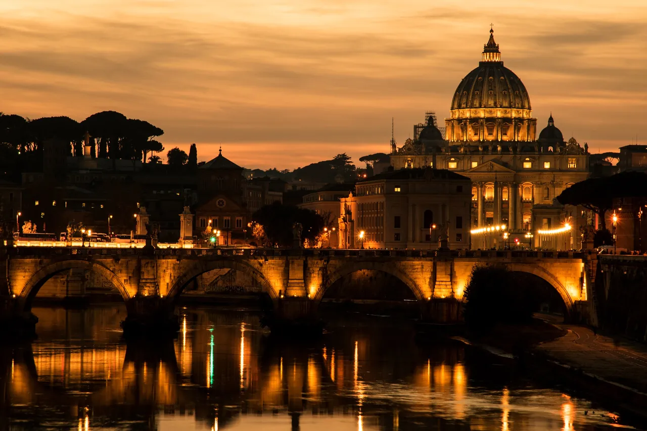St Peter's Basilica floodlit gold at dusk above the Tiber, its lights reflected in the water beneath the Ponte Sant'Angelo, under a deep amber sky.