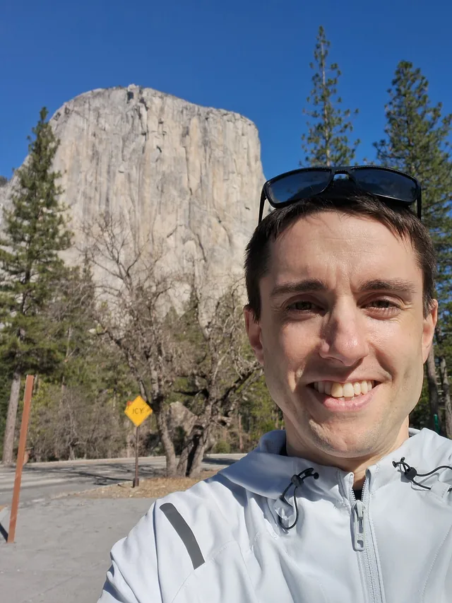 Jeff smiling in a light jacket on a sunny road in Yosemite Valley, with the granite face of El Capitan rising behind him against a deep blue sky.