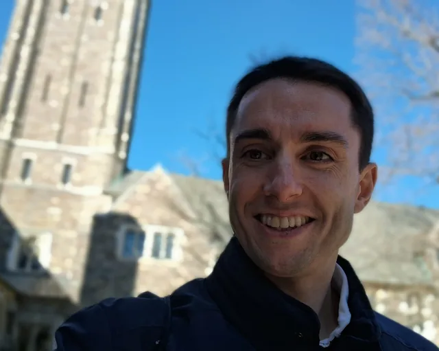 Jeff smiling in a dark jacket outdoors on a bright winter day, a stone collegiate tower rising behind him.